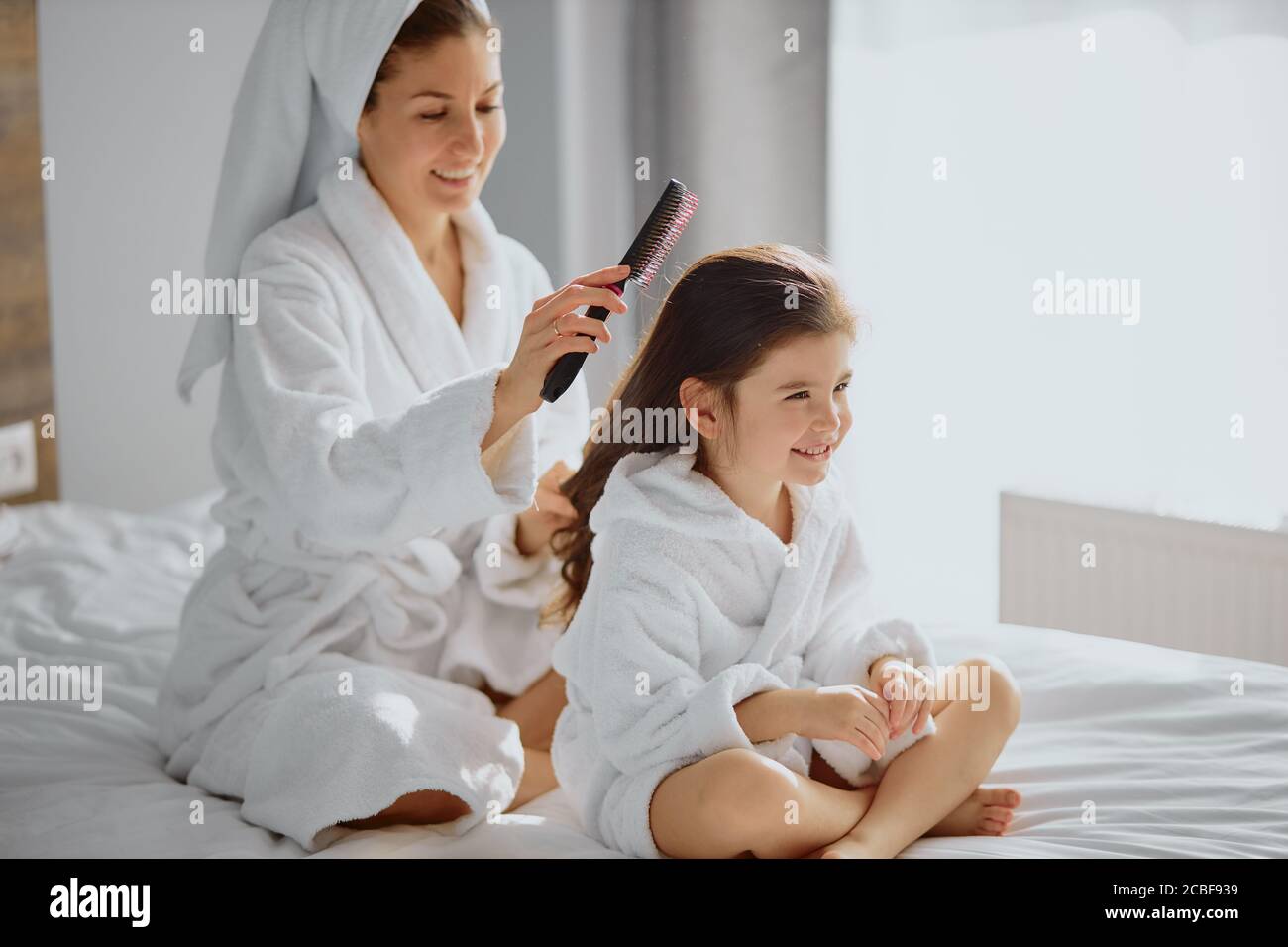 hermosa mujer peinando el pelo a su hija después de la ducha, sentada en la  cama en un dormitorio ligero, usando toalla y albornoz Fotografía de stock  - Alamy
