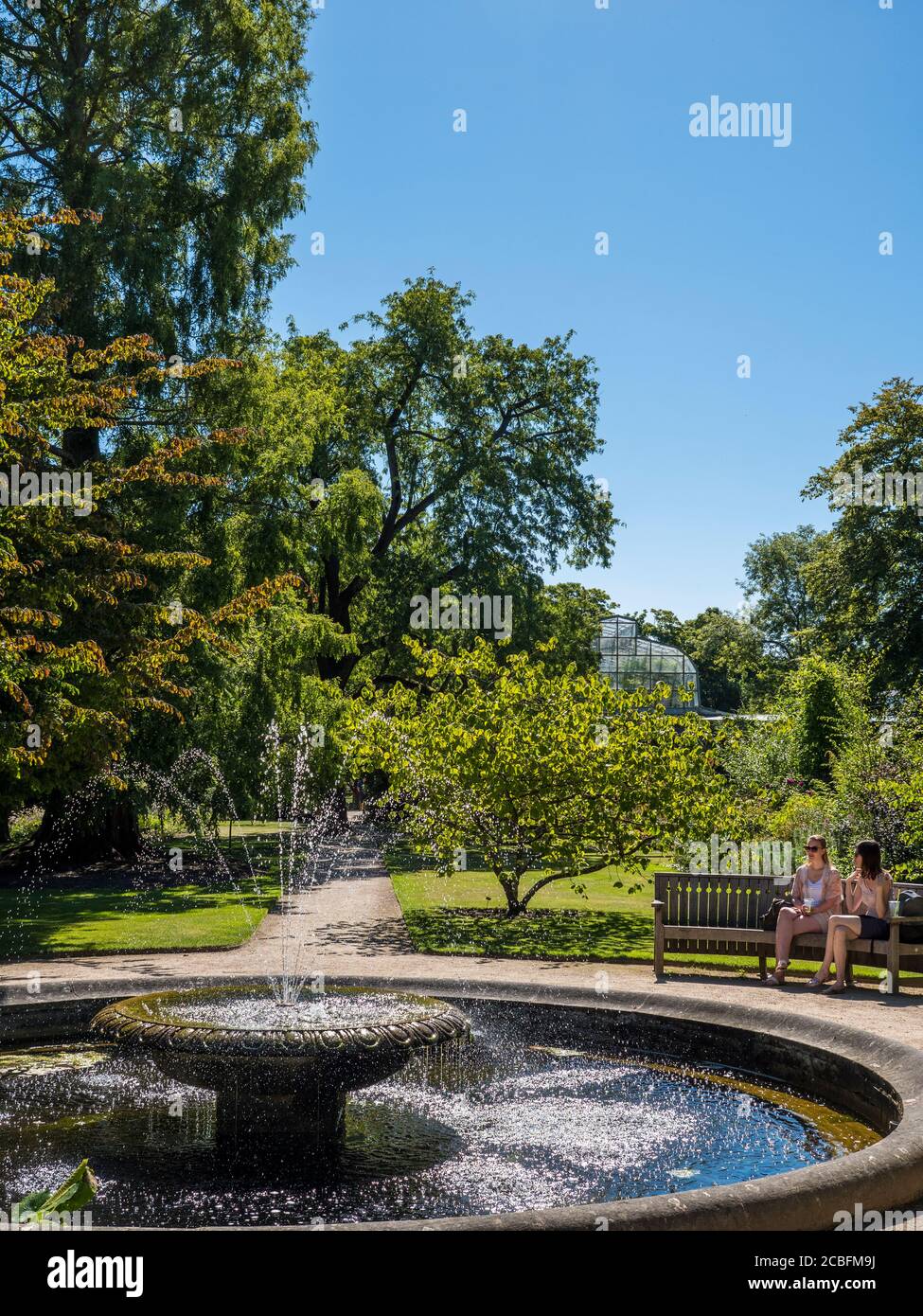 Dos mujeres jóvenes sentadas junto a la piscina con fuente, Universidad de Oxford Botanical Gardens, Oxford, Oxfordshire, Inglaterra, Reino Unido, GB. Foto de stock