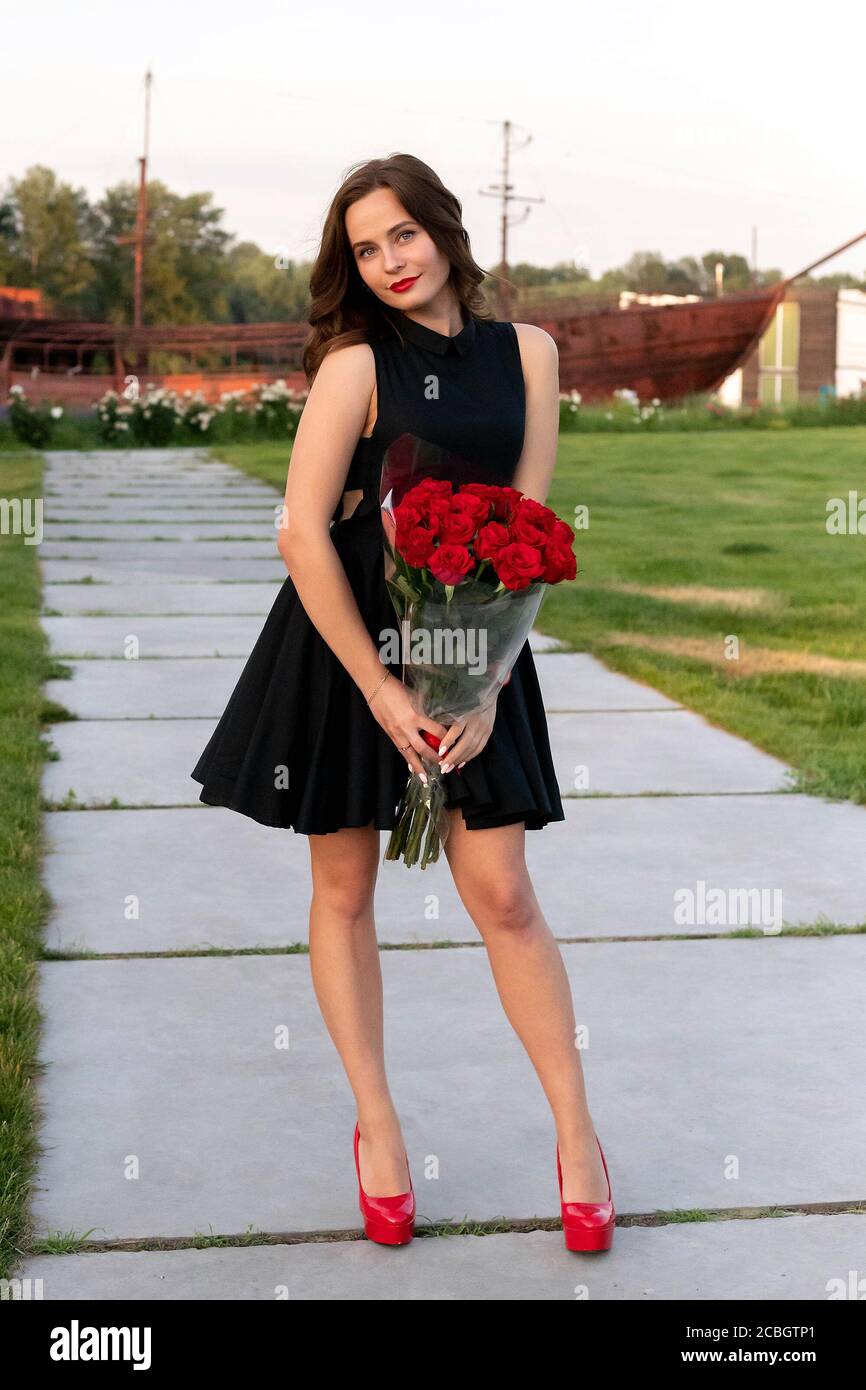 Atractiva niña de cumpleaños con pelo marrón en vestido negro posando, sonriendo y sosteniendo un ramo de rosas rojas sobre el fondo de barco de madera a Foto de stock