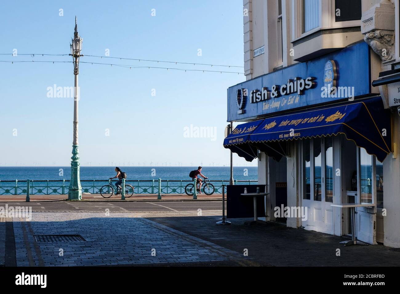 Pescaderías y patatas fritas en el paseo marítimo de Brighton durante el cierre. Día de verano con la tienda en el lado izquierdo, el mar y dos ciclistas en el fondo. Foto de stock