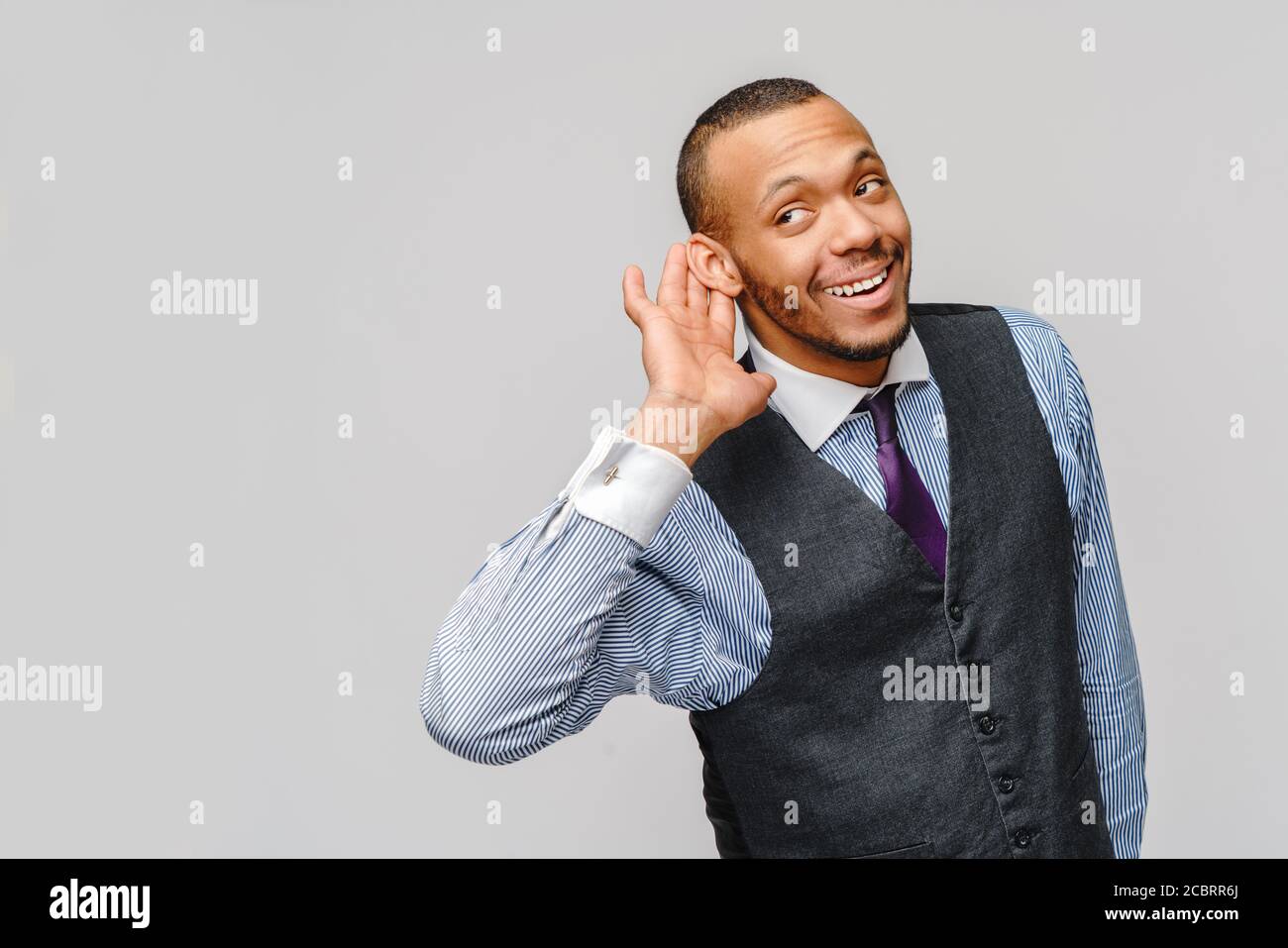 Joven hombre de negocios afroamericano que escucha algo o escucha, no puede oírle entender sobre fondo gris Foto de stock
