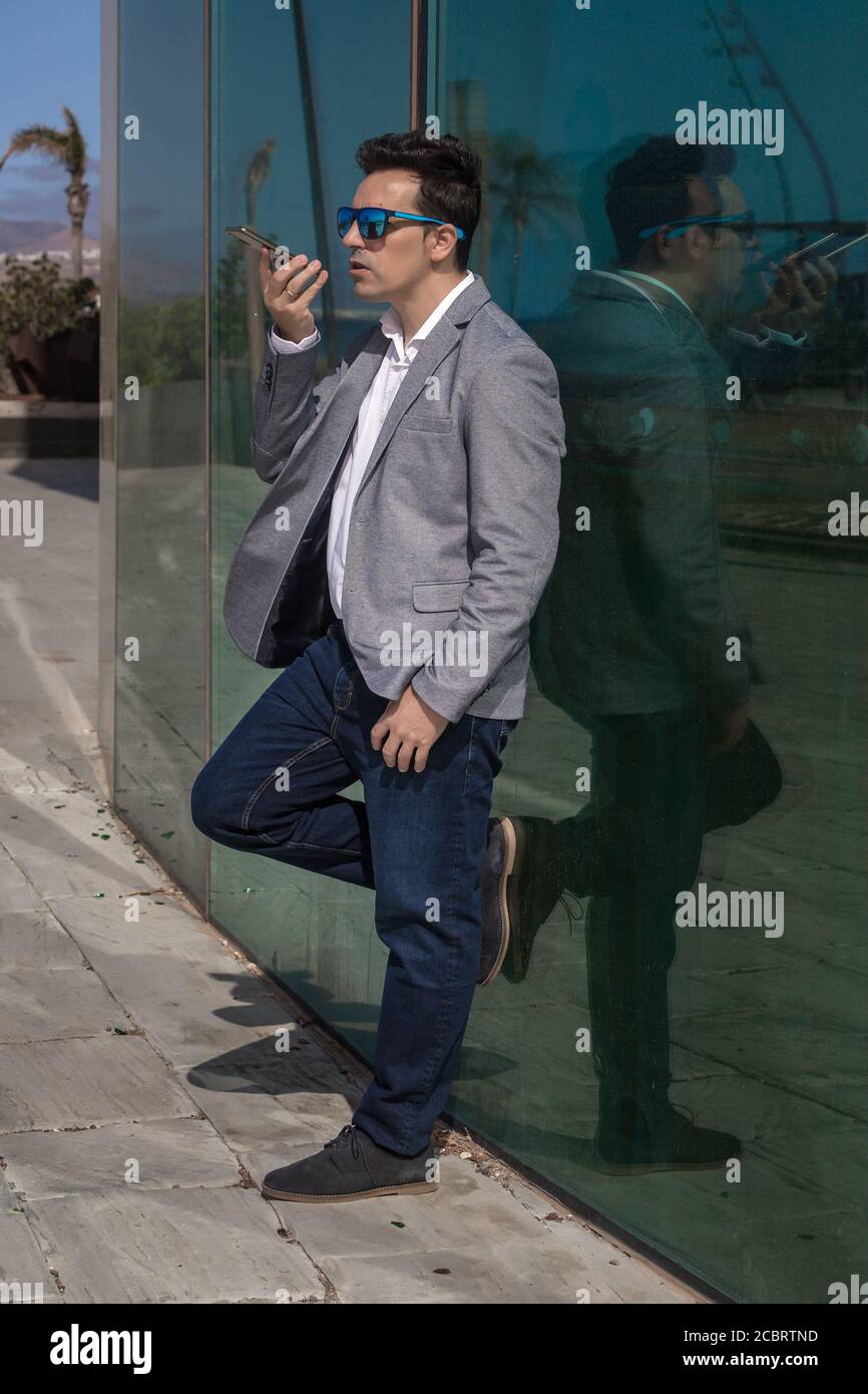 Hombre emprendedor de larga duración en ropa informal elegante inclinada en una pared de cristal y grabando un mensaje de audio Foto de stock