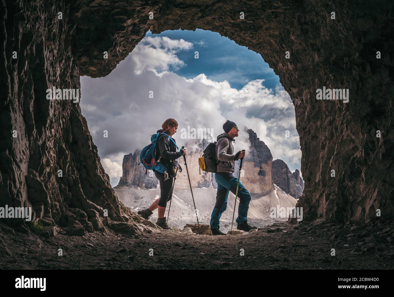 Un par de excursionistas caminando, con los picos de Tre Cime di Levarado en el fondo, vista desde una ventana de cueva, en Dolomitas, Italia. Foto de stock