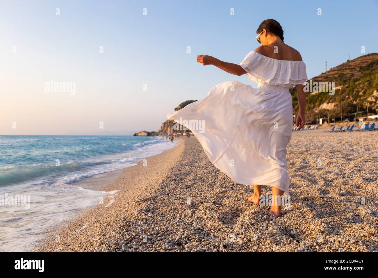 Una chica con vestido blanco caminando descalzo en la playa de Kathisma al atardecer, Lefkada, Islas Jónicas, Grecia Foto de stock