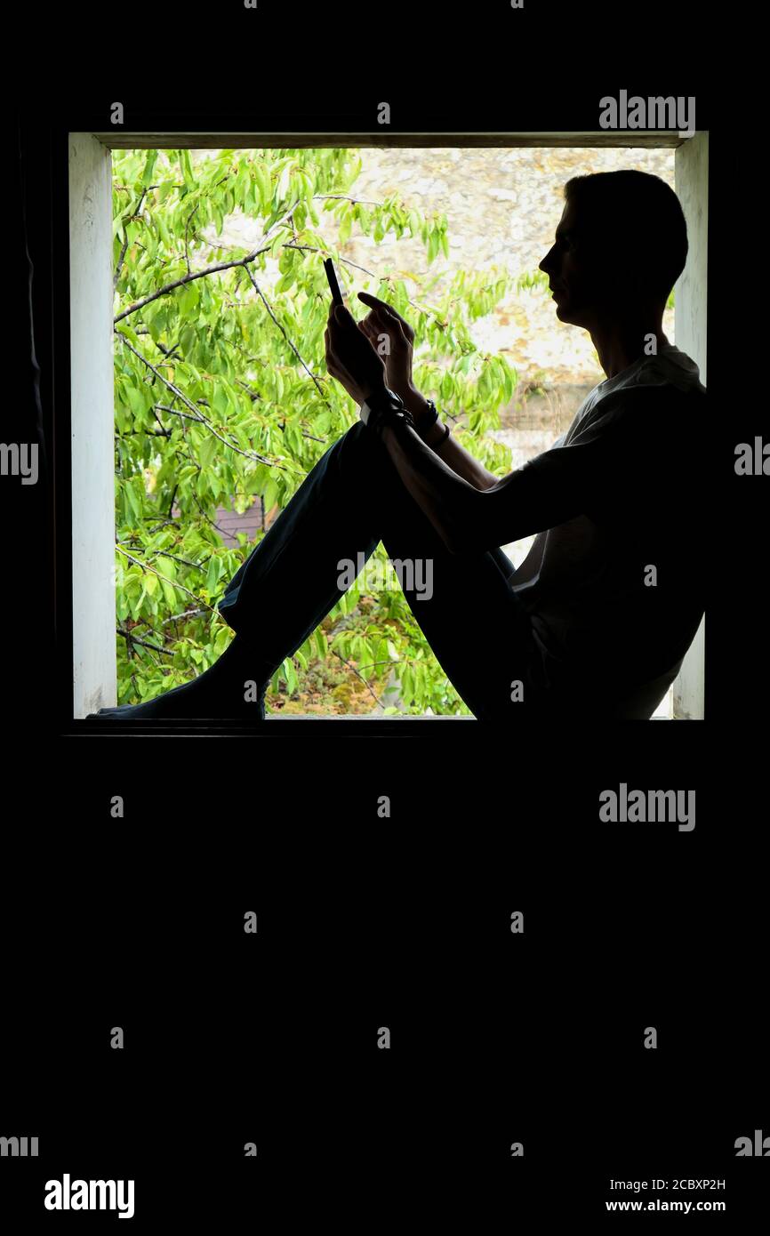 Persona que utiliza tecnología. Silueta de un hombre en el interior de una ventana, con un smartphone en sus manos. Foto de stock
