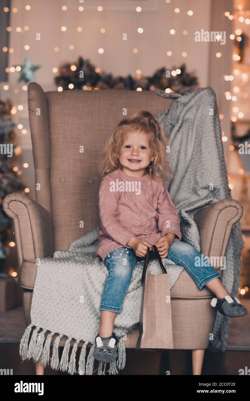 Lindo bebé divertido niña de 3-4 años de edad con pantalones vaqueros y  suéter de punto sentado en silla sobre luces brillantes de Navidad en la  habitación primer plano. Vacaciones de invierno