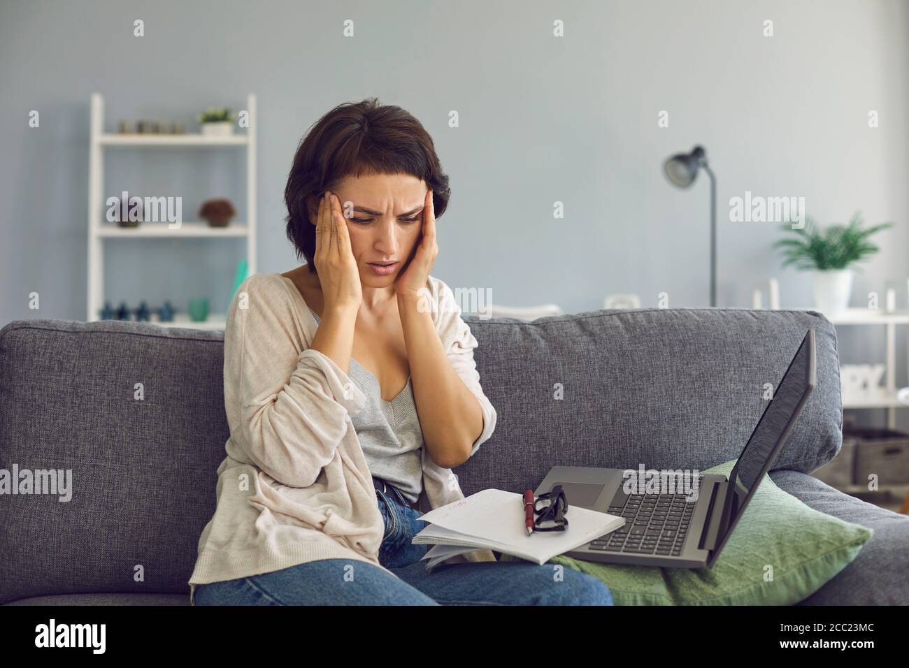 El freelancer sobretrabajado que sufre de dolor de cabeza cerca de la computadora portátil en casa. Mujer estresada con agotamiento profesional en el interior Foto de stock