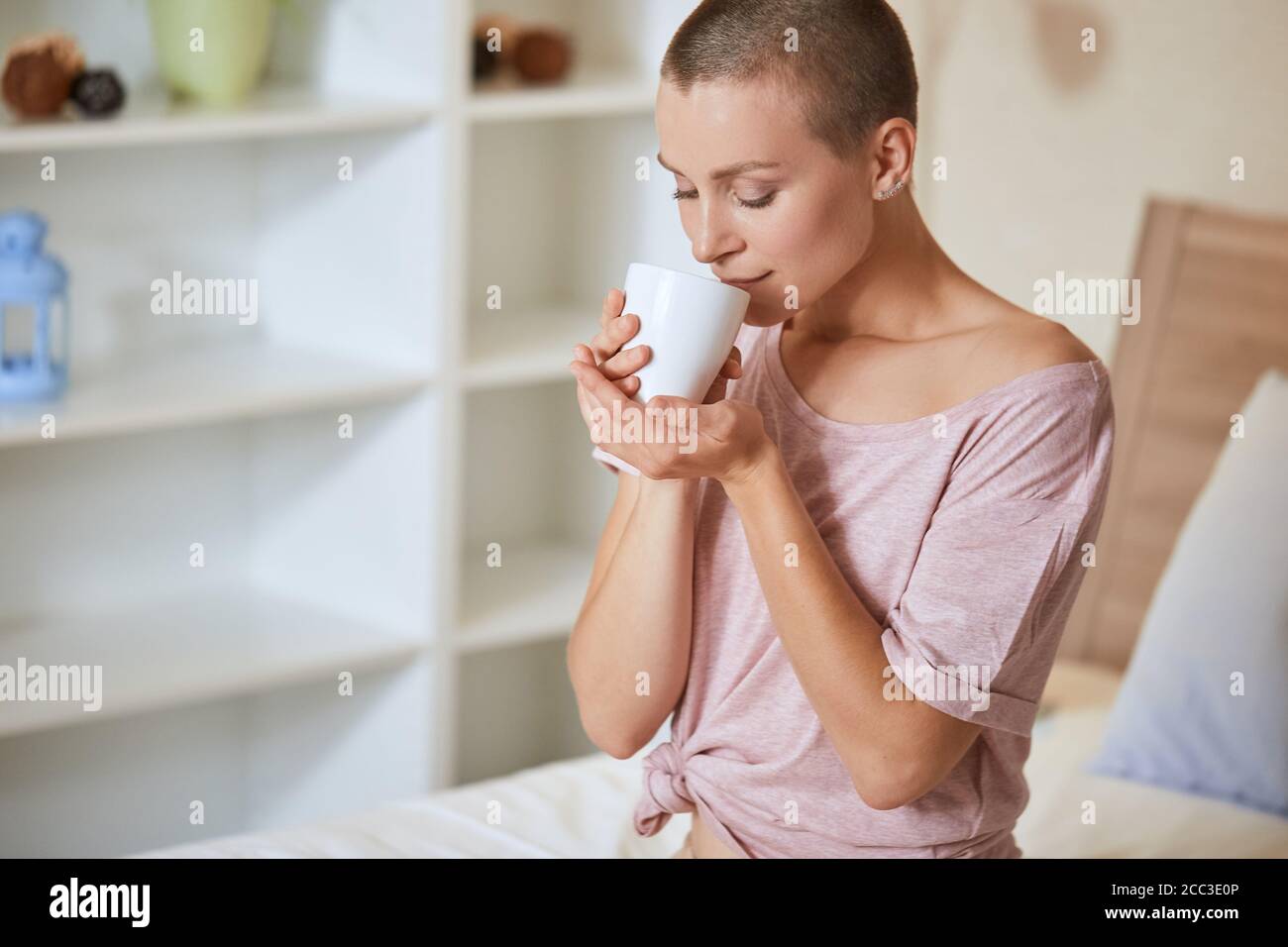 Hermosa joven con pelo corto sentarse en la cama en la habitación de luz y disfrutar de beber té o café por la mañana en casa. Foto de stock