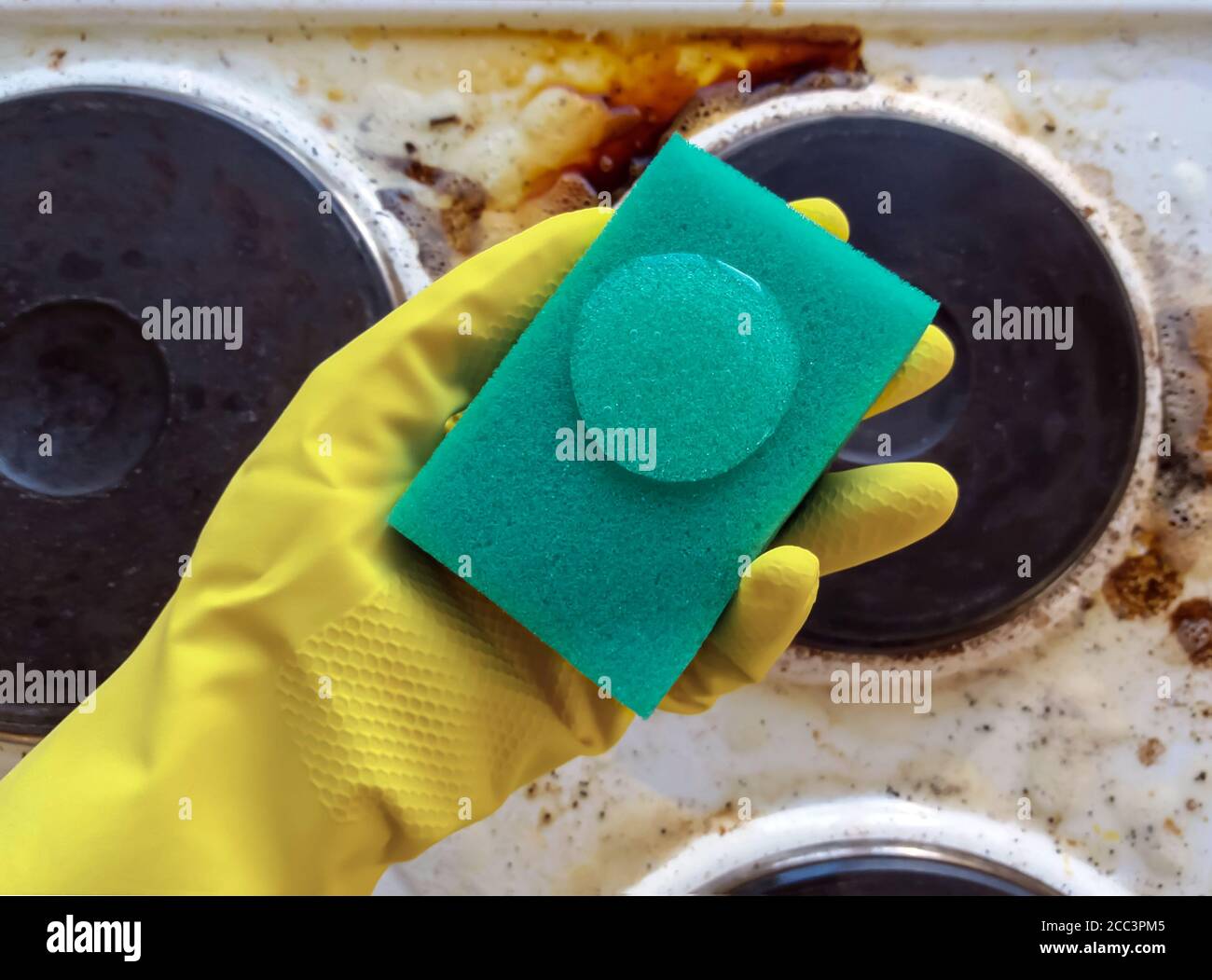 mano en el guante de la casa sosteniendo la esponja con el líquido de limpieza encima estufa sucia Foto de stock