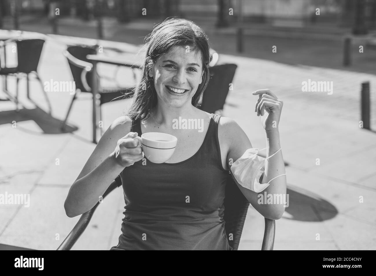 Mujer joven sentada en una cafetería con protector quirúrgico mascarilla facial en el brazo mientras bebe café al aire libre Escenarios públicos en el Coronav Foto de stock