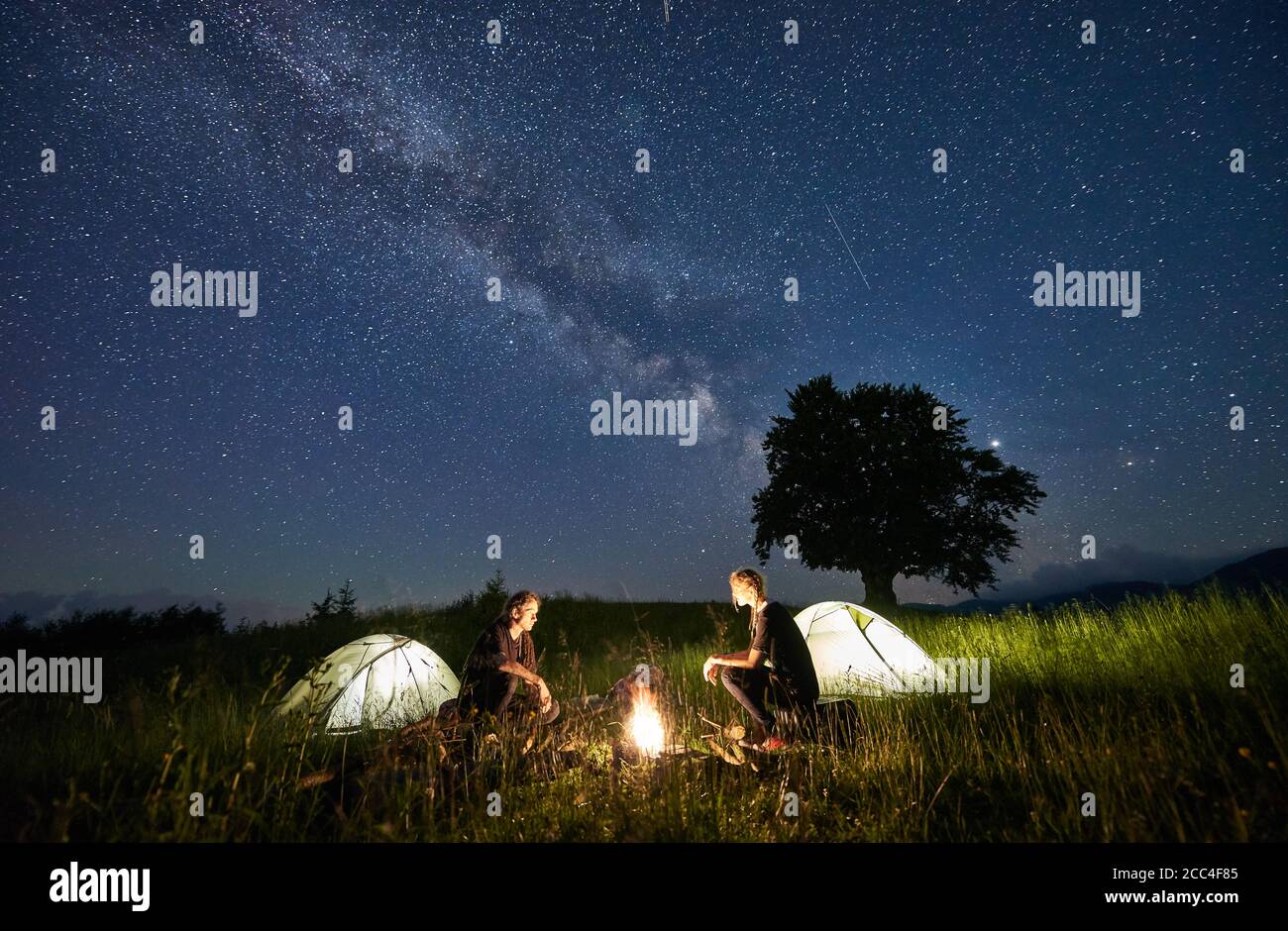 Hermosa vista del cielo estrellado nocturno sobre la pradera con  excursionistas cerca de carpas de campamento iluminadas. Turistas sentados  cerca de fogatas bajo el mágico cielo azul con estrellas. Concepto de viaje,