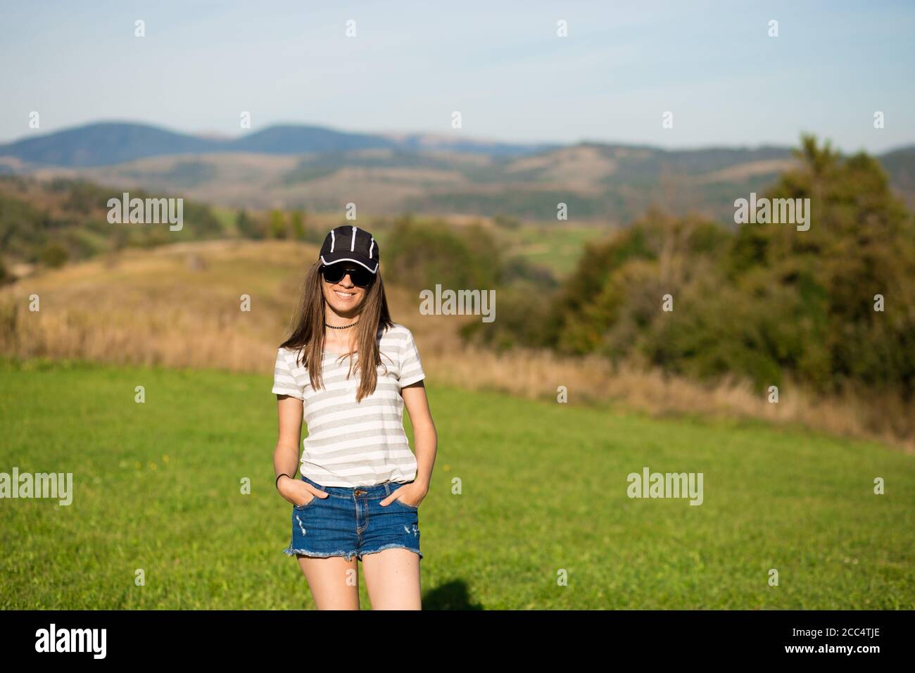 Mujer despreocupada feliz en gafas de sol, gorra y pantalones cortos  pantalones cortos caminar en verde pradera de hierba en la cima de la  montaña disfrutando de la naturaleza. Libertad. Disfrute. Daydreamin
