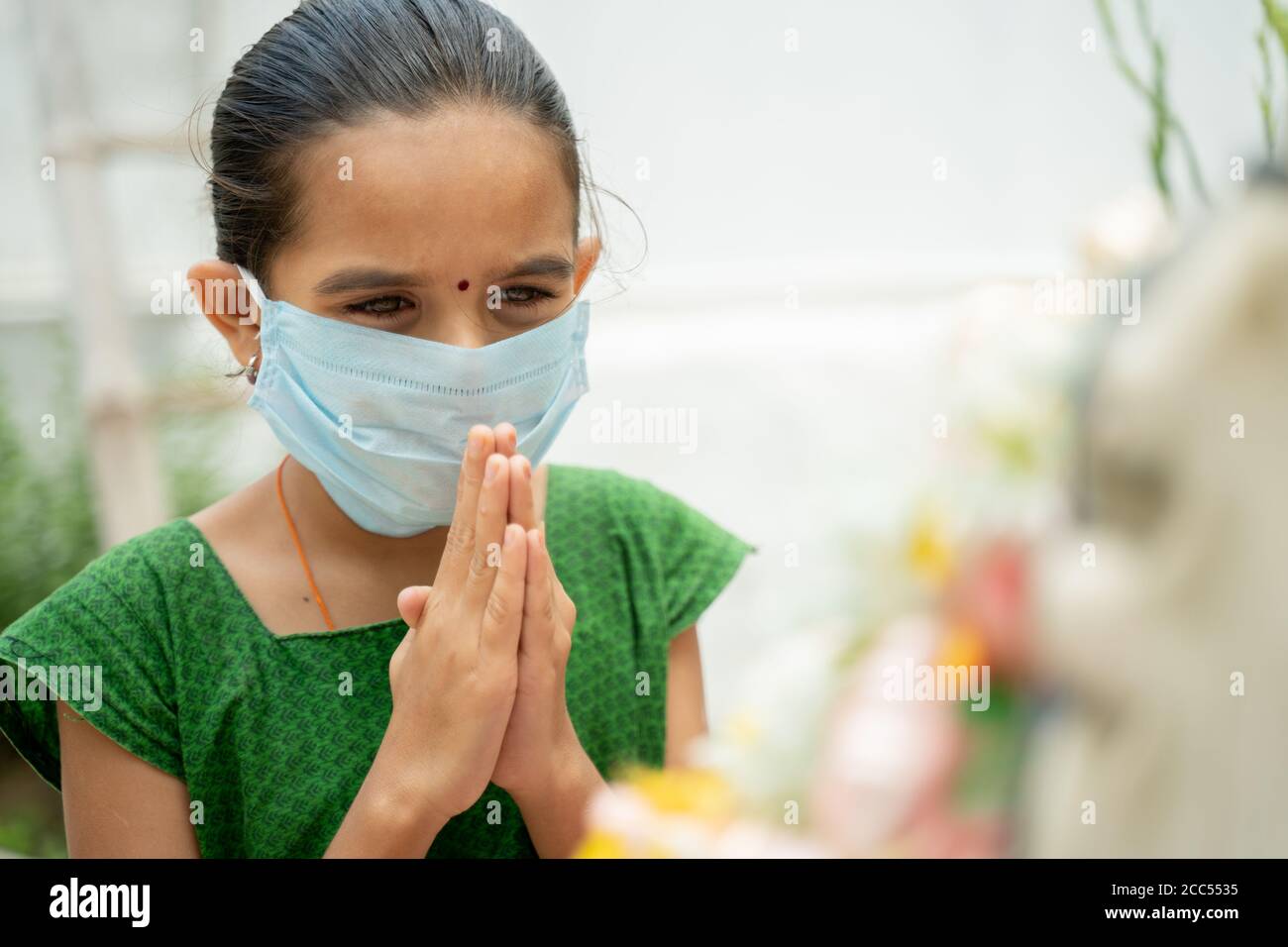 Niña joven en máscara médica rezando a dios para protegerse del coronavirus o covid-19 doblando las manos en el gesto de namaste. Foto de stock