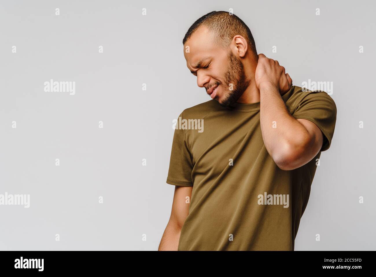 joven afroamericano con camiseta verde sobre fondo gris siente malestar físico para el dolor de cuello masajeando los músculos tensos Foto de stock