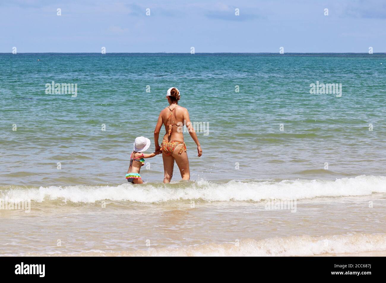Mamá joven que lleva al niño alegre al mar. Mujer delgada en bikini con una pequeña hija chapoteando en las olas en una playa de arena, el concepto de vacaciones de verano Foto de stock