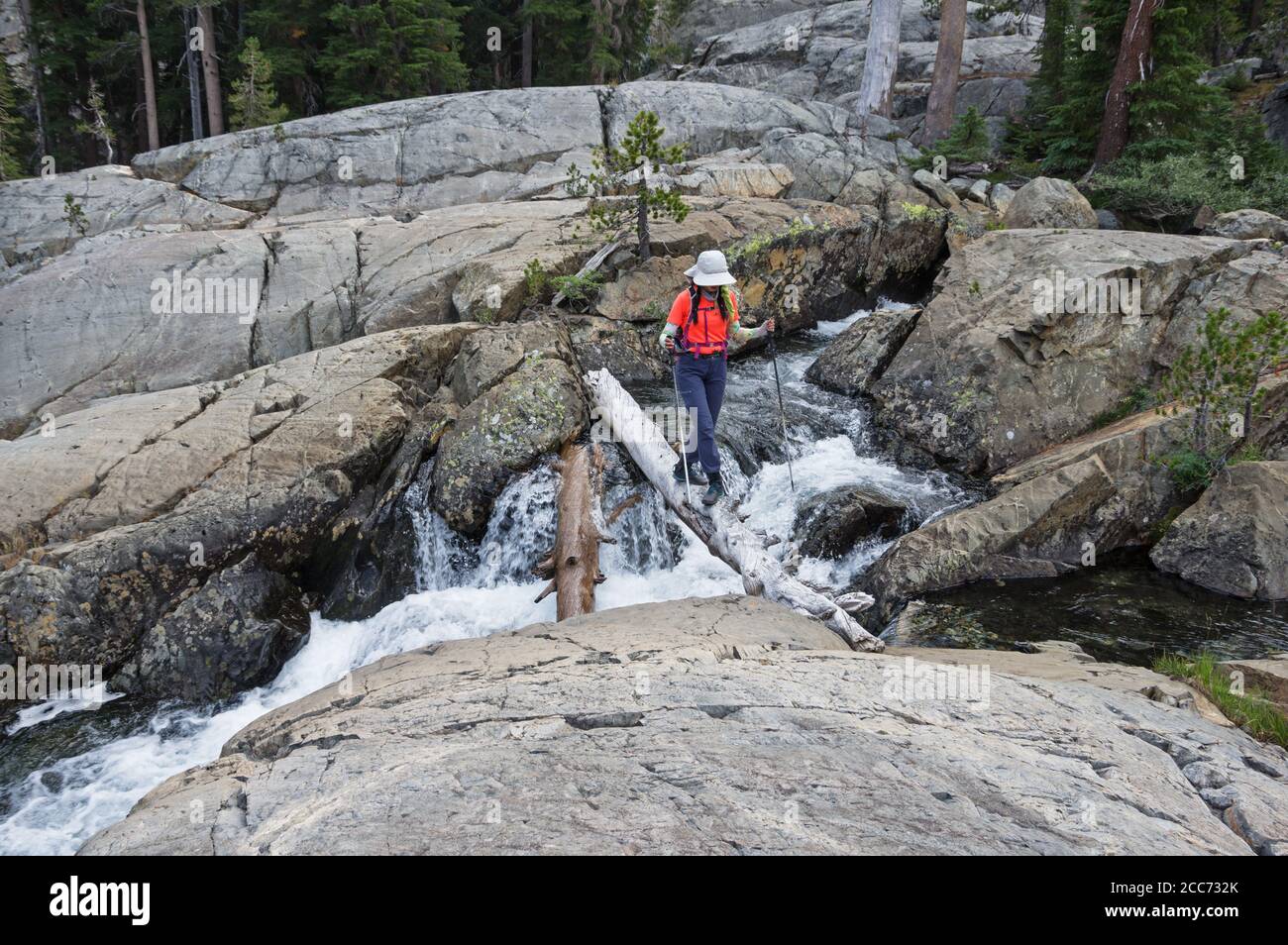 mujer caminante cruzando un arroyo a través de rocas pulidas glaciales en un registro Foto de stock