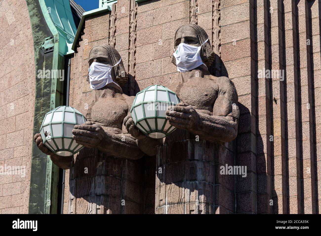 Lámpara esférica que sostiene estatuas con máscaras de cara por la entrada principal de la Estación Central de Ferrocarril durante el brote de coronavirus en Helsinki, Finlandia Foto de stock