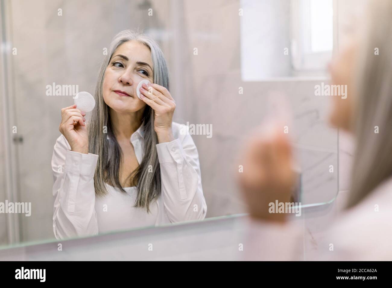 Concepto de cuidado e higiene de la piel. Encantadora señora mayor de pelo gris usando una almohadilla de algodón con agua micelar para eliminar el relleno de la cara, de pie Foto de stock
