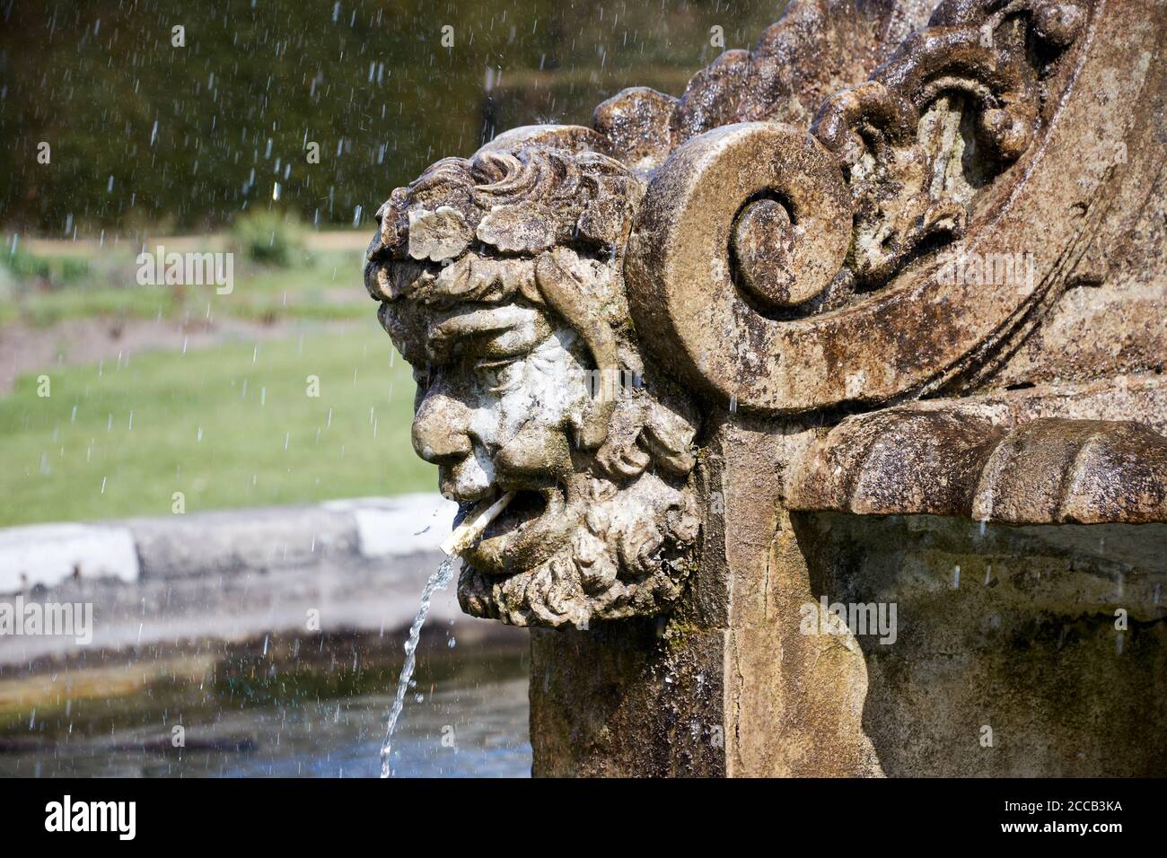Fuente de piedra en la forma de una cabeza de mans barbuda con el agua que se precipena de la boca en un jardín inglés formal, la escultura Foto de stock