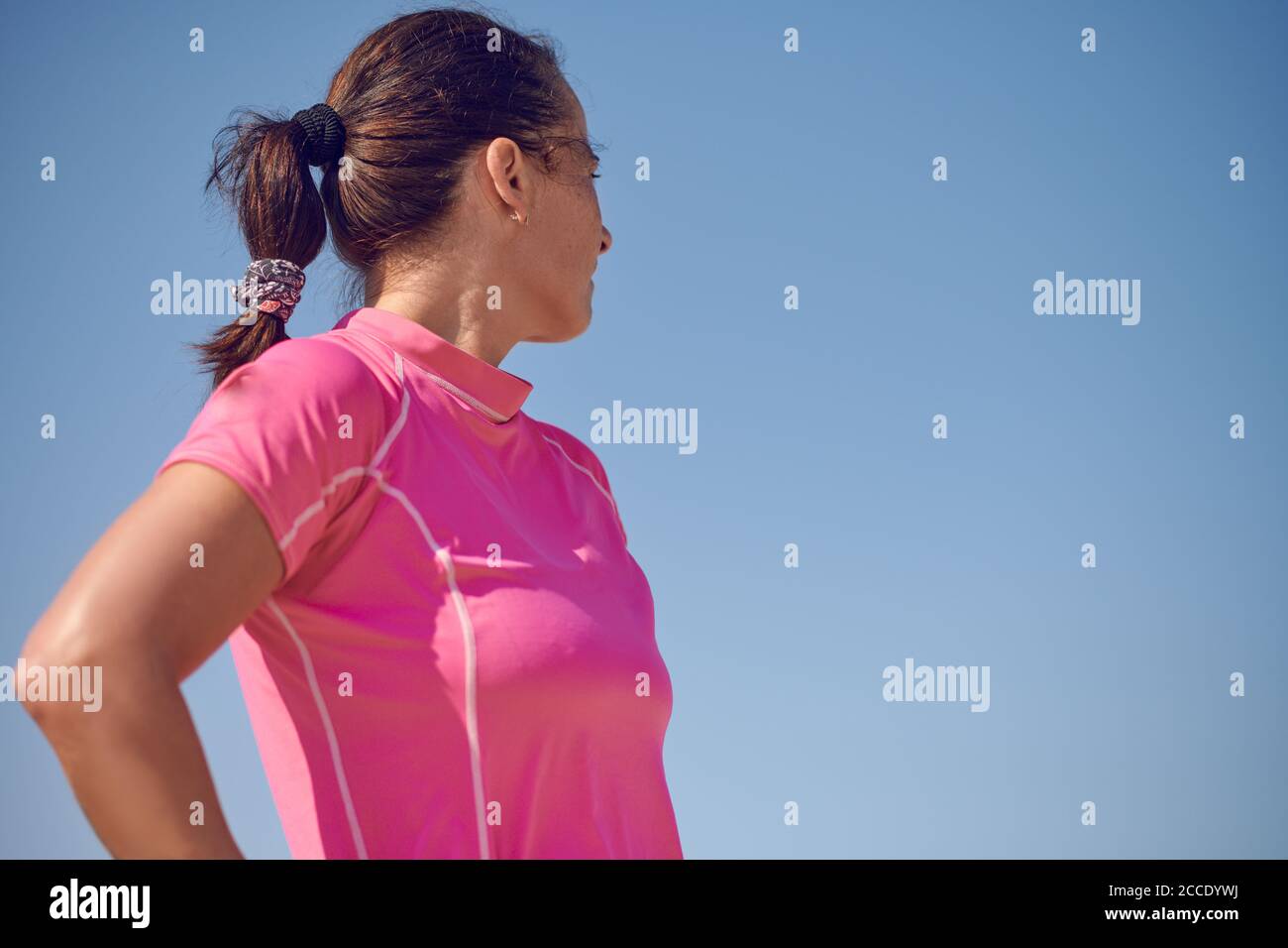 Mujer deportiva con cola de caballo en una parte superior rosa mirando hacia atrás sobre su hombro en una vista de ángulo bajo contra un cielo azul claro y soleado Foto de stock