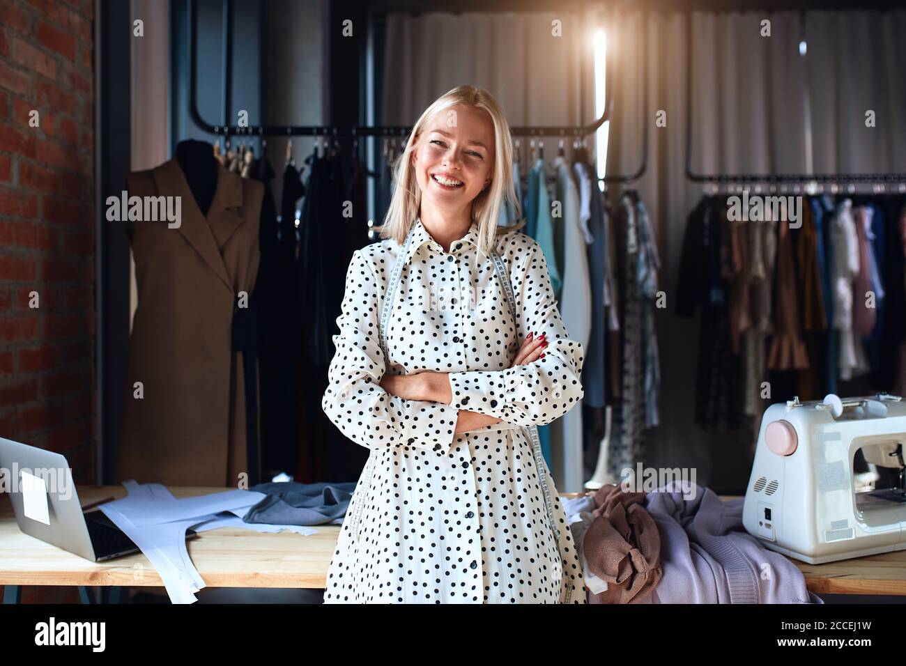 Hermoso puesto de diseñador de moda caucásico y felizmente sonrisa en el lugar de trabajo, mirar la cámara. Vestido con vestido blanco. Estudio de diseño de fondo Foto de stock