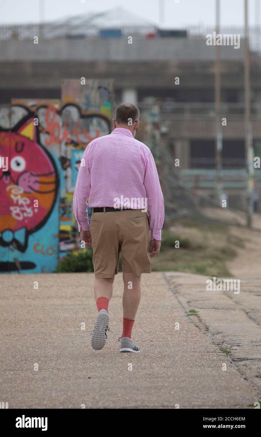 Un hombre en una camisa rosa, pantalones cortos y largos caminando a lo largo del paseo marítimo en Brighton, East Sussex, Reino Unido Fotografía de stock -