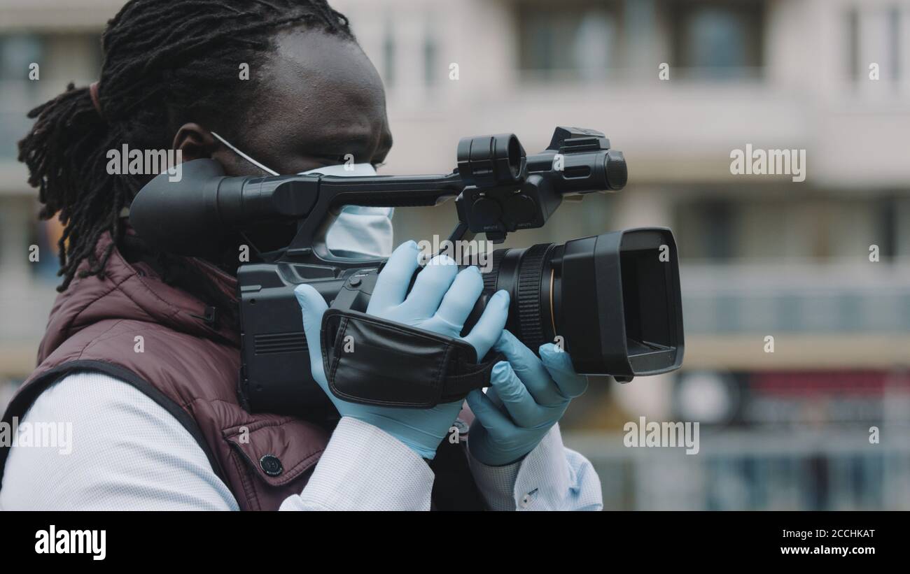 De cerca, videógrafo negro africano con máscara medikal mirando a través del visor en la cámara de vídeo . Foto de alta calidad Foto de stock