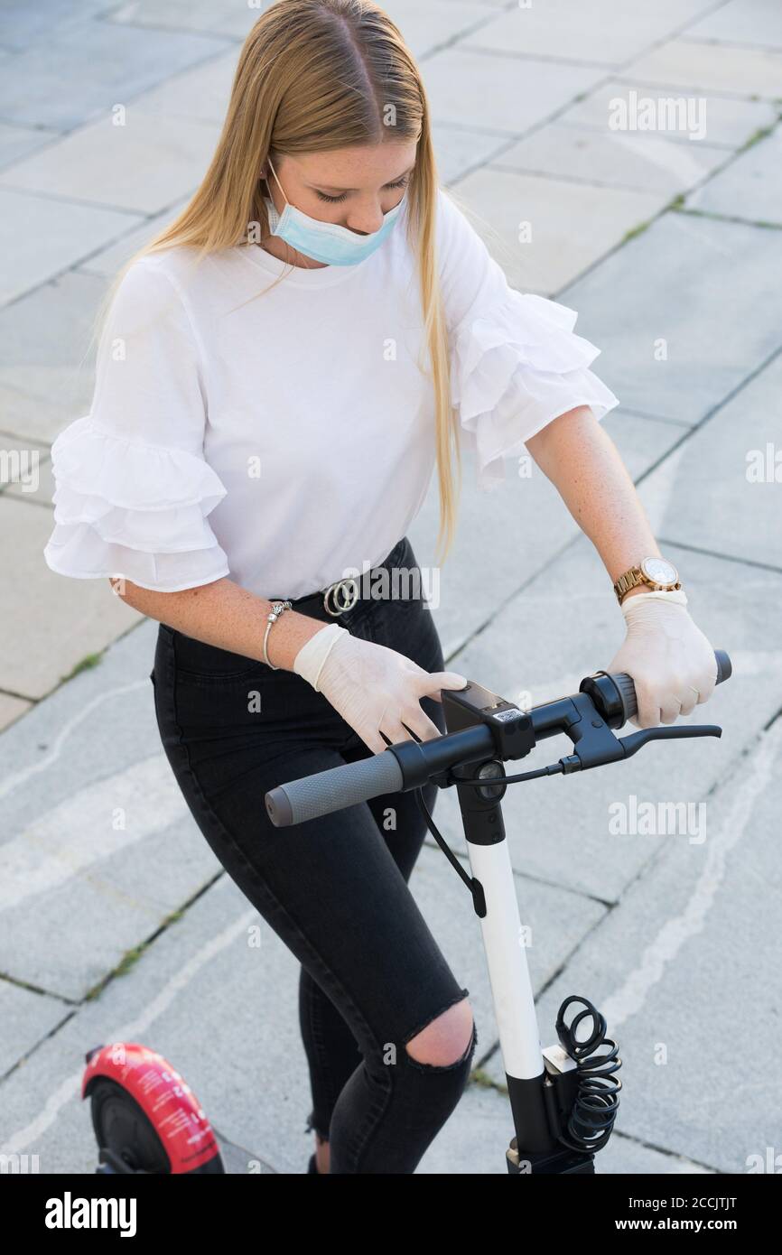 Niña de moda fashinable usando corona virus protector máscara facial y guantes de goma mientras que utiliza scooters eléctricos de alquiler en el entorno de la ciudad. Eco Foto de stock