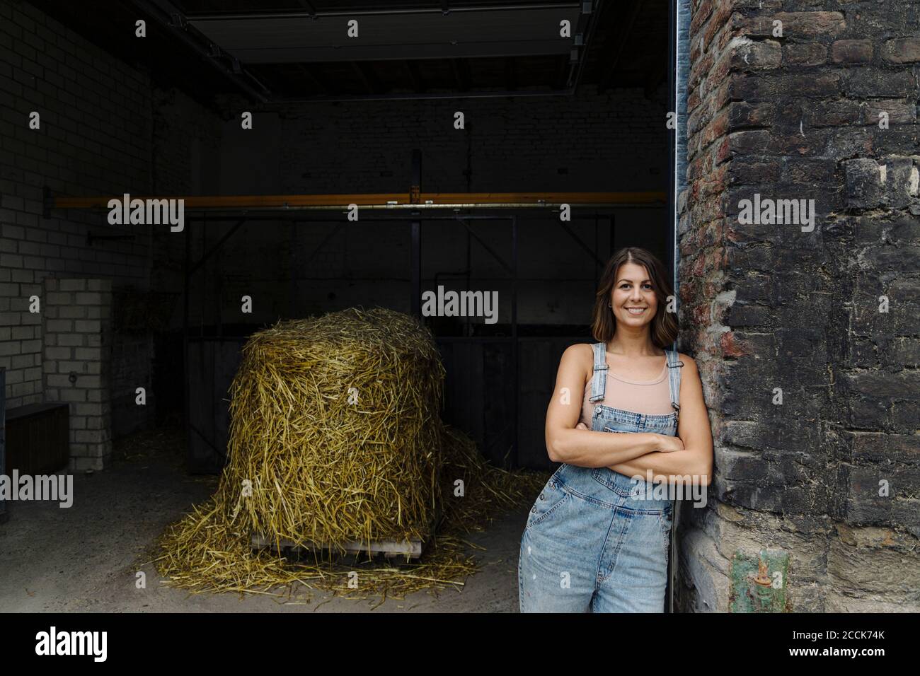 Retrato de una joven sonriente apoyada contra una pared de ladrillo en un granero en una granja Foto de stock