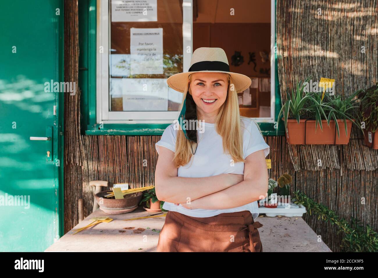 Mujer sonriente propietaria con sombrero con los brazos cruzados de pie mesa fuera del vivero de la planta Foto de stock
