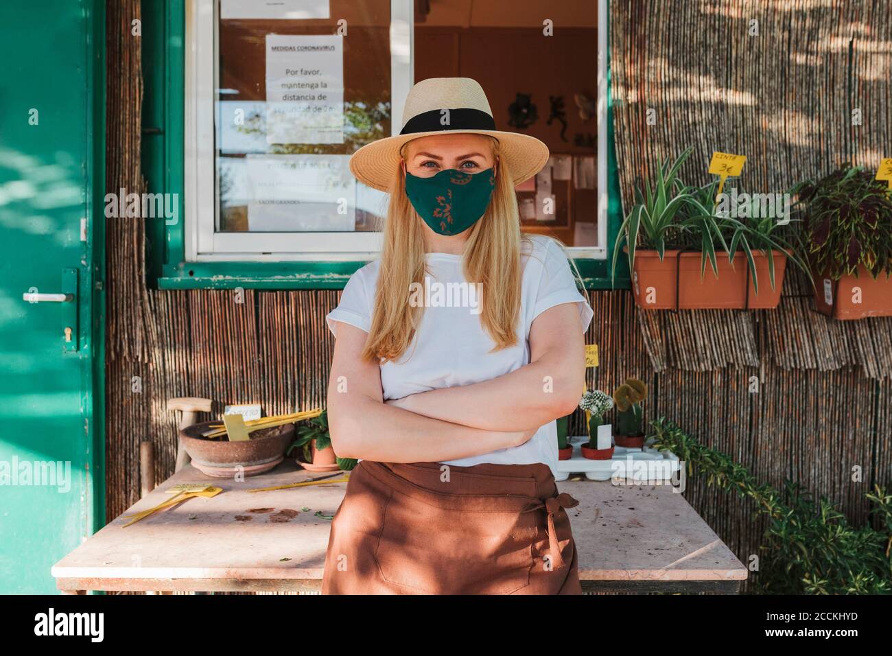 Mujer propietaria con sombrero y máscara con los brazos cruzados de pie por mesa fuera del vivero de plantas Foto de stock