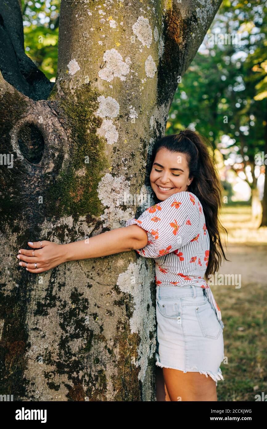 Sonriente joven con los ojos cerrados abrazando el tronco del árbol mientras de pie en el parque Foto de stock