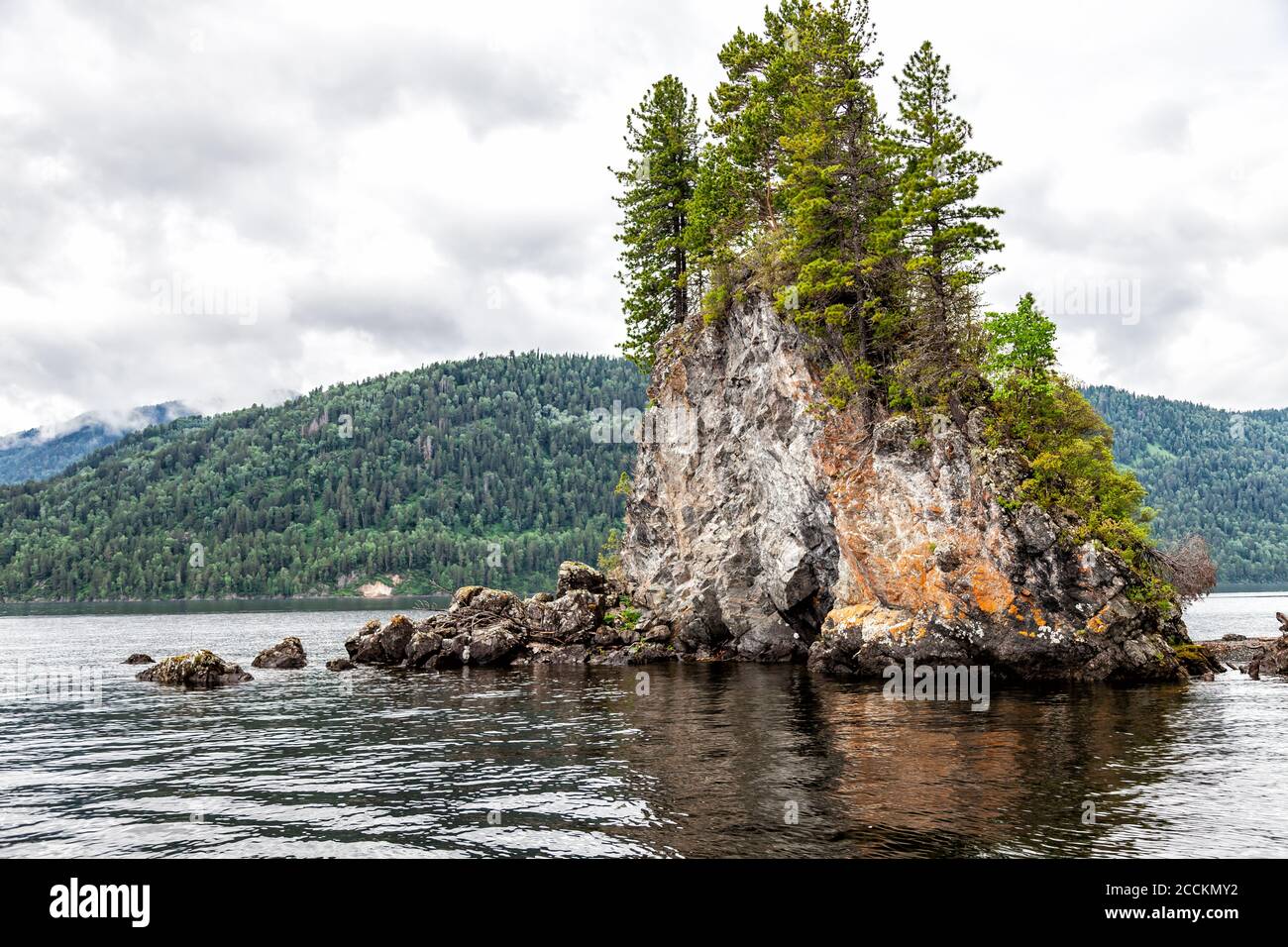 Una isla rocosa con árboles en crecimiento en el Piedras en el medio del lago Teletskoye en Altai sobre el fondo de las montañas en el nublado Foto de stock