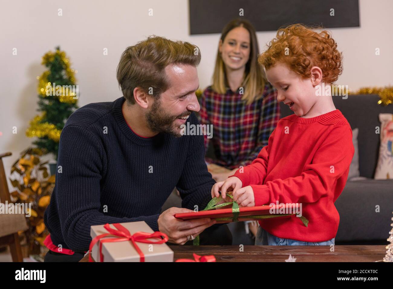 Mujer sonriente mirando al padre dando regalo de Navidad al hijo en la sala de estar Foto de stock