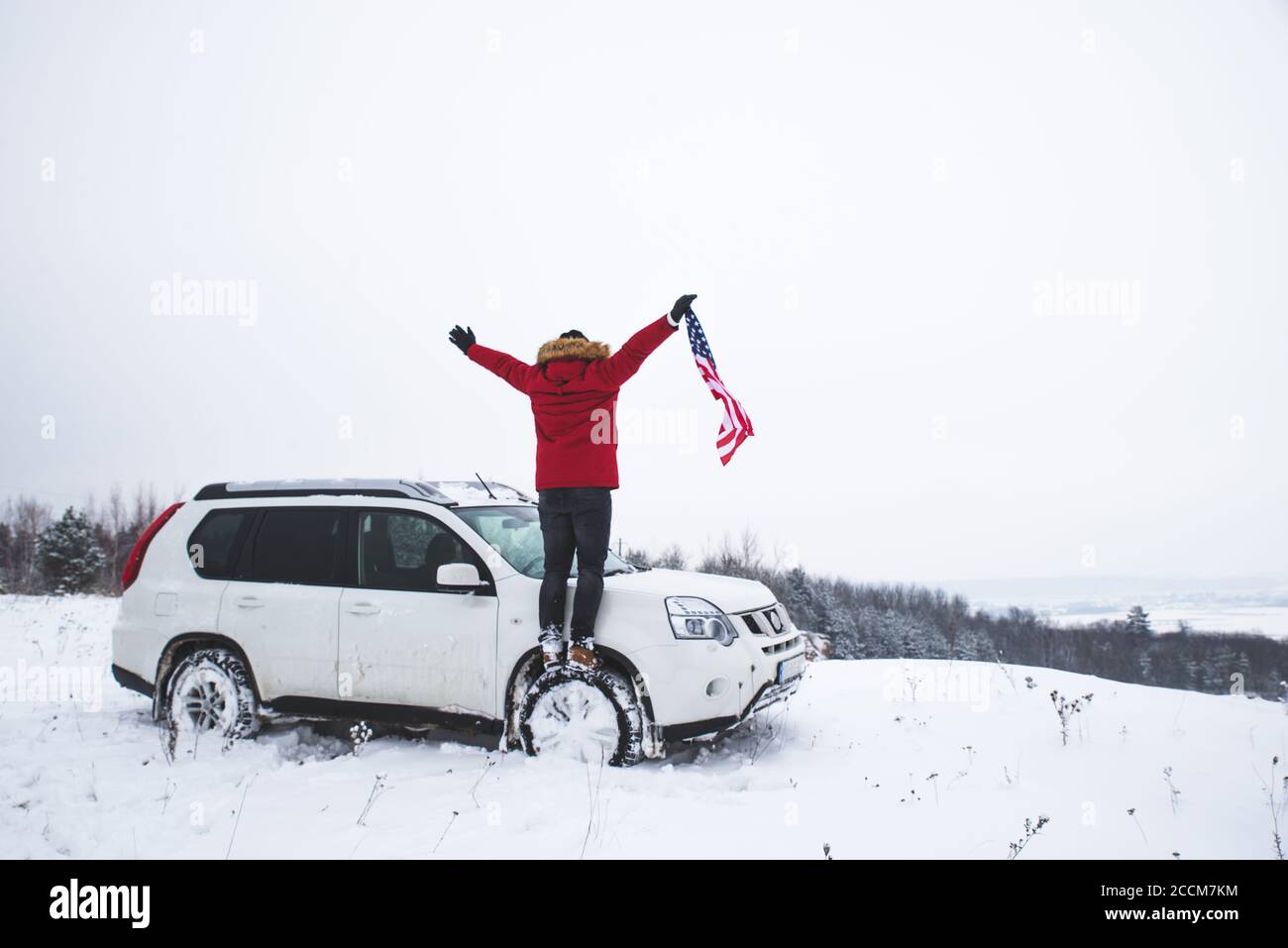 Hombre de pie en la cima de la colina con la bandera de EE.UU. en sus manos cerca de suv coche buscando hermosa vista de invierno Foto de stock
