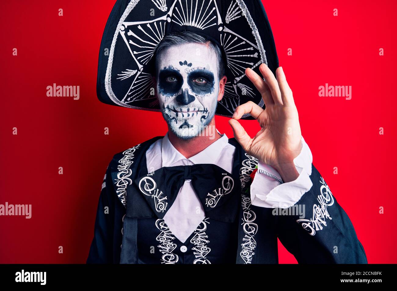 Hombre joven con el día mexicano del traje muerto sobre rojo sonriendo positivo haciendo ok signo con la mano y los dedos. Expresión exitosa. Foto de stock