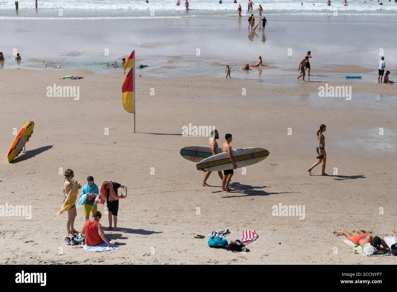 Día de la playa: Dos jóvenes llevando tablas de surf, familia, niños jugando, gente caminando, tabla de rescate de surf. Playa principal, Byron Bay, Australia Foto de stock