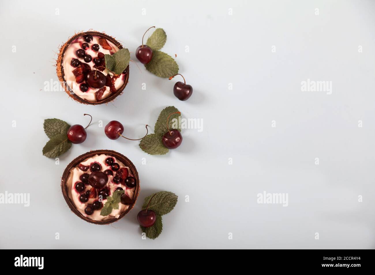 Postre crema batida con cerezas y grosellas y hojas de menta en mitades de coco sobre un plato blanco. Postre de keto de verano Copiar spaes. Lay plano. Foto de stock