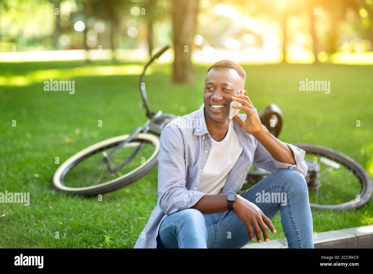 Atractivo hombre negro sentado en el suelo cerca de su bicicleta y.. hablando por teléfono celular en el parque Foto de stock