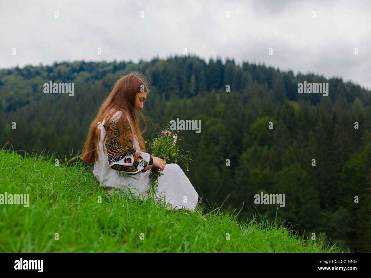 niña con pelo suelto en un bordado con un ramo de flores silvestres se sienta sobre hierba verde contra el fondo de las montañas de los cárpatos Foto de stock