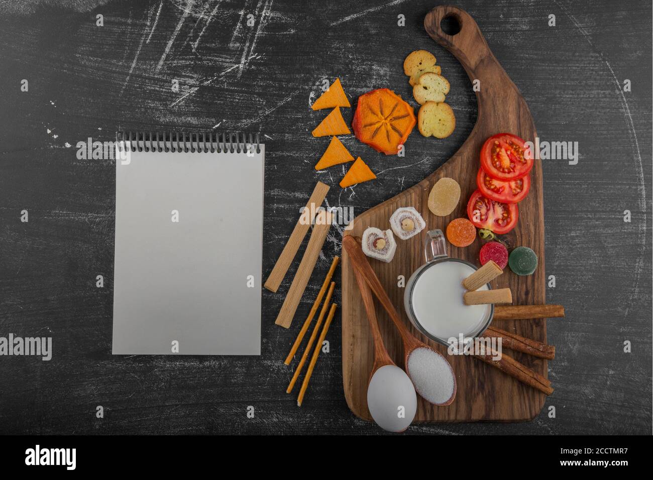 Tabla de aperitivos con patatas fritas, galletas saladas y pasteles en el plato de madera con un libro de recibos aparte Foto de stock