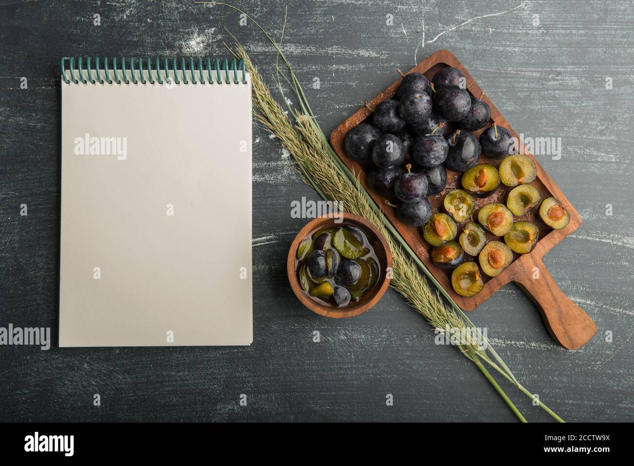 Ciruelas negras enteras y cortadas en una tabla de madera con un libro de recibos aparte Foto de stock