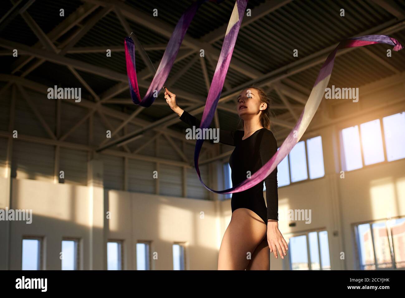 Profesional talentoso femenino de arte gimnasta agitando la mano con cinta violeta, trabajando en estudio brillante, tiro inferior, concepto de estilo de vida saludable Foto de stock