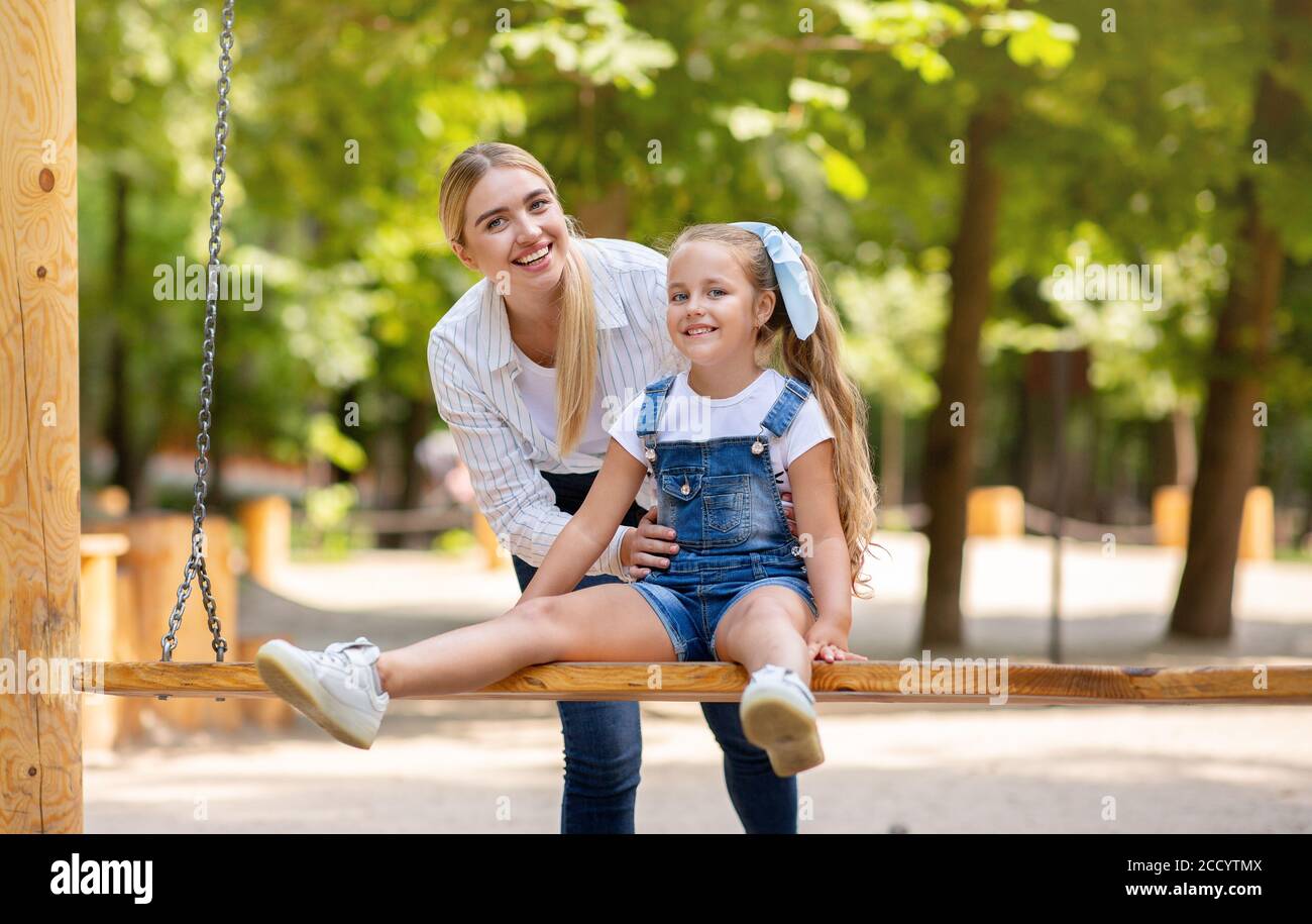 Madre feliz empujando a la hija pequeña en columpios en el patio de juegos al aire libre Foto de stock