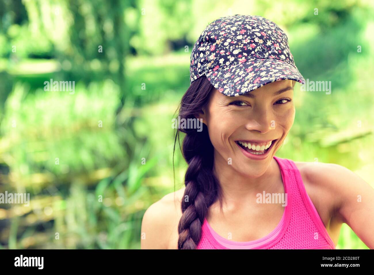 Joven feliz chica asiática sana sonriendo a la cámara. Mujer multirracial caucásica China con gorro de moda y trenza de pelo en el verde parque de verano Foto de stock