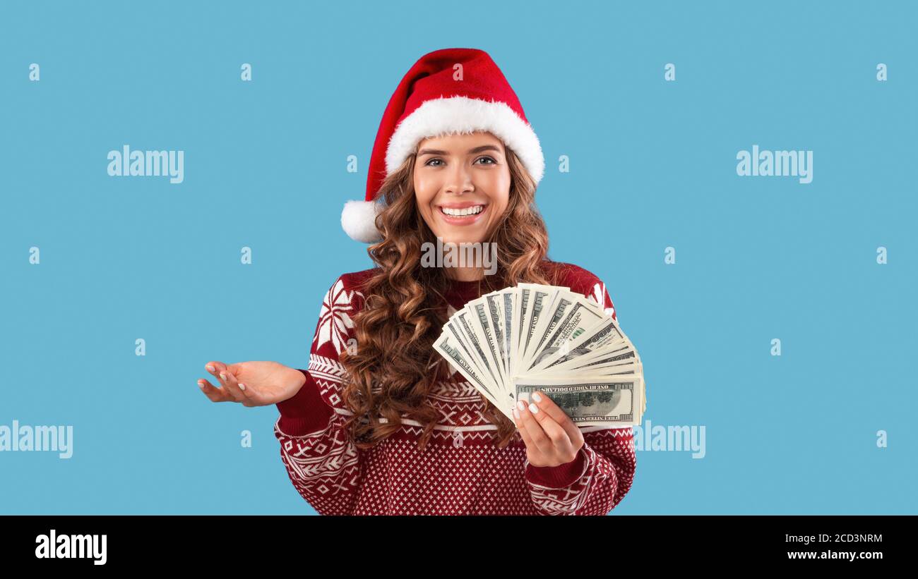 Concepto de descuentos navideños. Feliz milenial chica en el sombrero de Santa y suéter sosteniendo fan de dinero sobre fondo azul Foto de stock