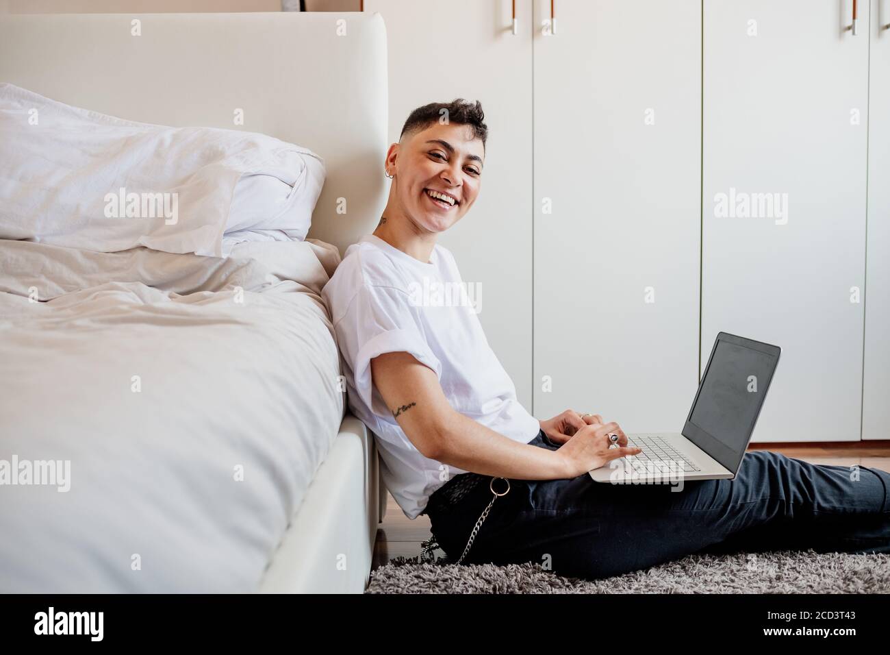 Mujer joven con cabeza afeitada sentada en el dormitorio, usando el ordenador portátil, sonriendo con la cámara. Foto de stock