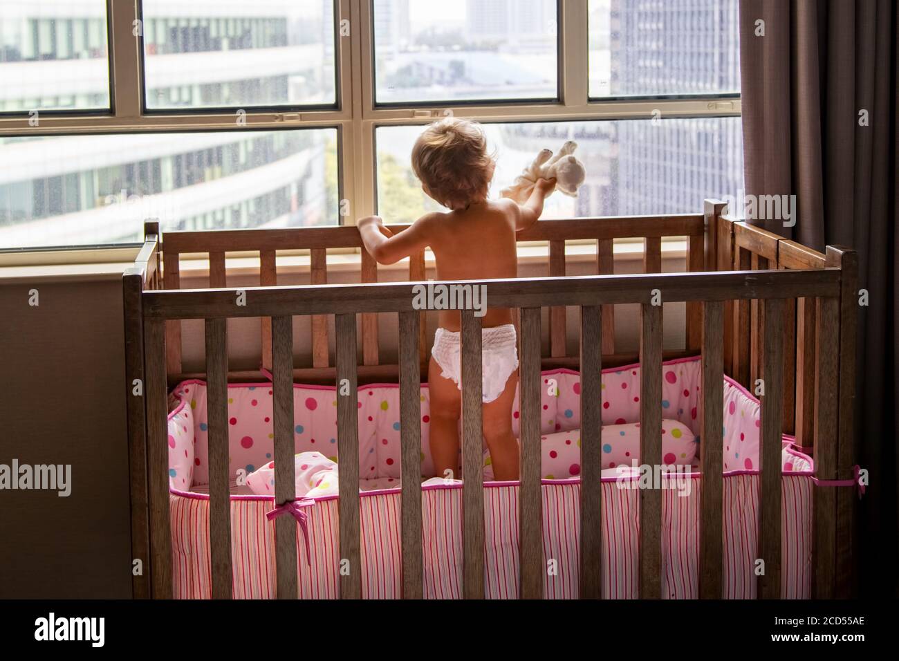 concepto de estancia en casa. niño pequeño con gato de juguete está parado en la cuna y mirando por la ventana a los rascacielos de la ciudad Foto de stock