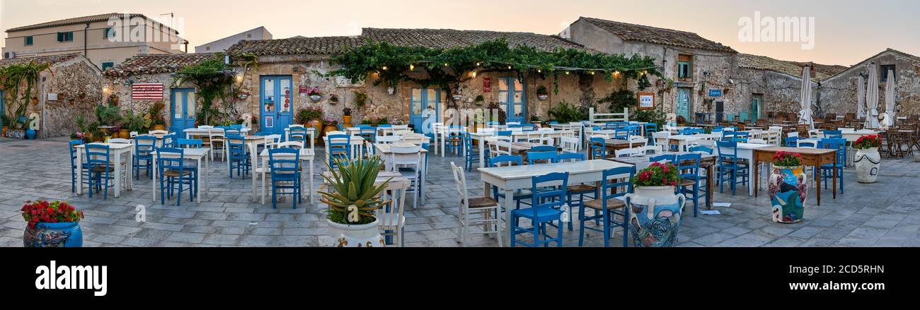 Mesas y sillas en restaurantes italianos tradicionales en la plaza principal del histórico pueblo Marzamemi, Siracusa, Sicilia durante un día soleado. Foto de stock