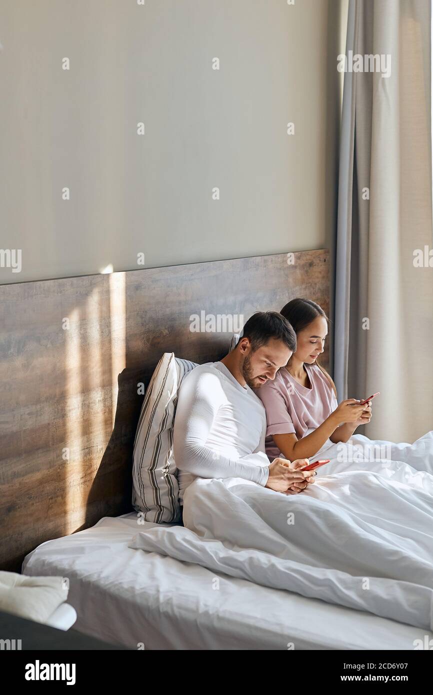 Jóvenes amantes agradables en pijamas tener tiempo feliz en la habitación  de hotel, sol brillando a través de una gran ventana, enviar mensajes de  texto con amigos usando teléfonos celulares, mirar lejos